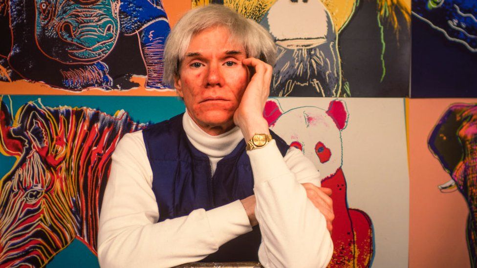 Andy Warhol ศิลปินป๊อปชาวอเมริกัน (1928 - 1987) นั่งหน้าภาพวาดหลายภาพใน 