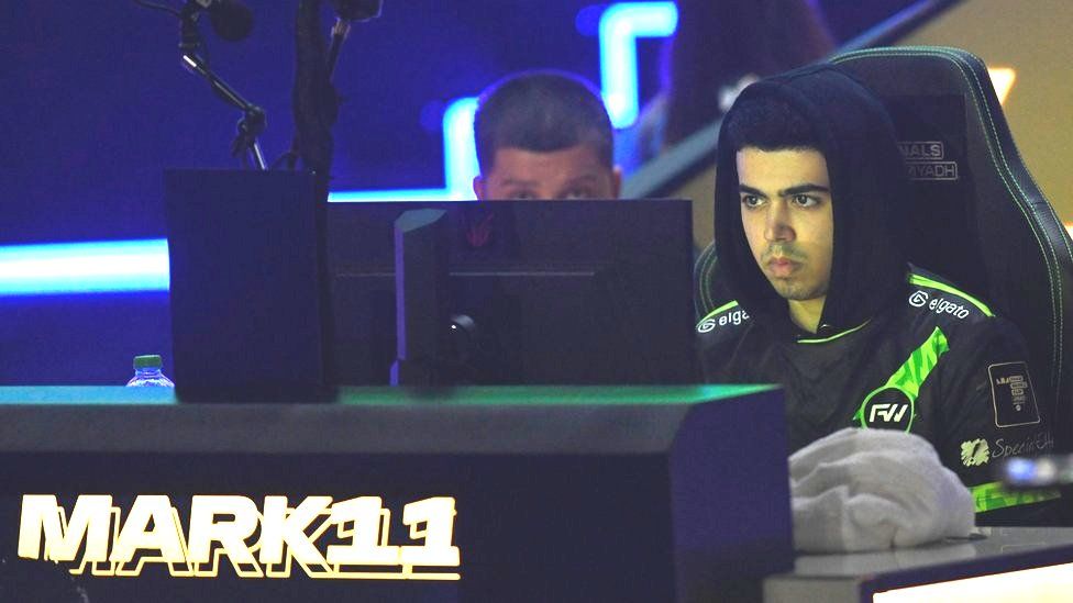 Mark Zakhary concentrates in a dimly lit arena as he competes in the FIFAe World Cup 2023. He's sat in a gaming chair, and mostly obscured by his monitor. He's wearing a black hoodie - with hood up - with lime green piping on the shoulders and a lime green slash across the chest. It says "elgato" in white on both shoulders. His face is tinged green from the monitor light. We can see his team-mate's head poking over the monitor, which sits on a desk with his screen name - Mark11 - written in gold, embossed block capitals