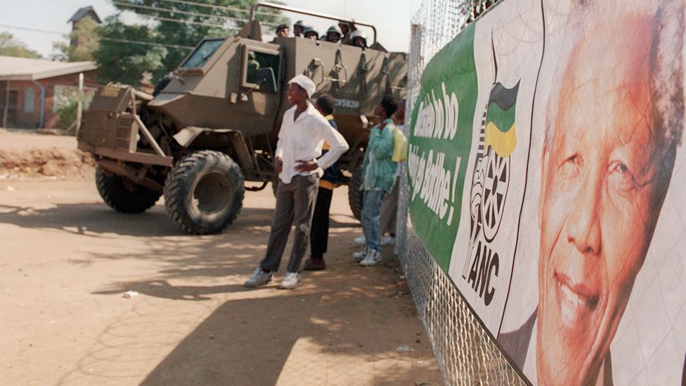 People next to an armoured vehicle and a poster of Nelson Mandela in South Africa - 25 April 2024