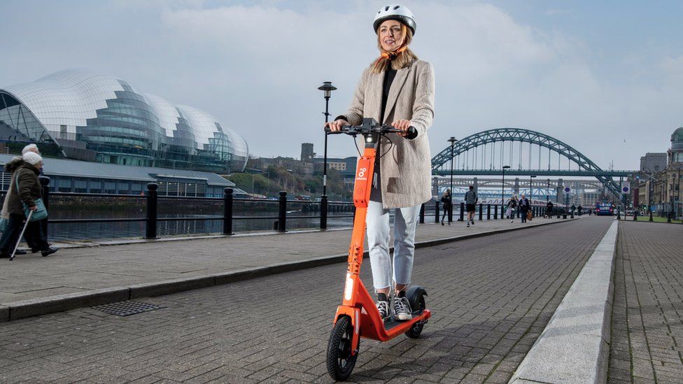 A woman pictured on one of the orange e-scooters on Newcastle Quayside with the Tyne Bridge and Sage music centre behind her.