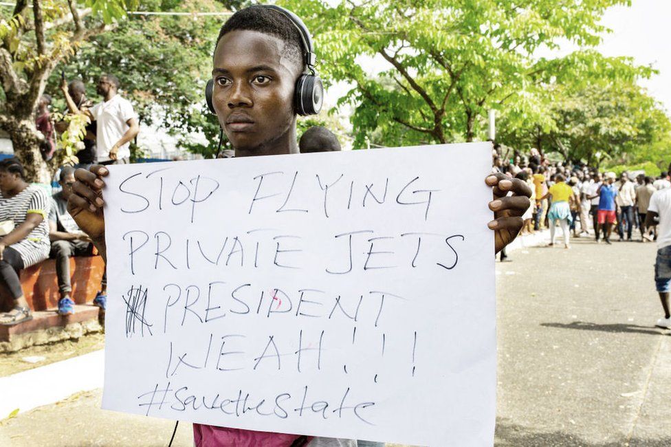A man holds a sign that says "stop flying private jets President Weah, #savethestate"