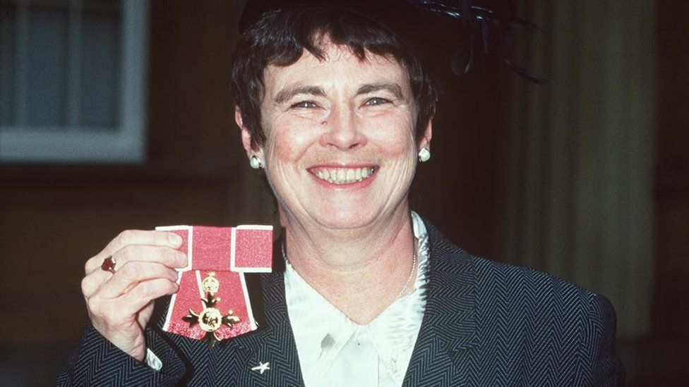 angela mason holds up her oBE while she smiles at the camera