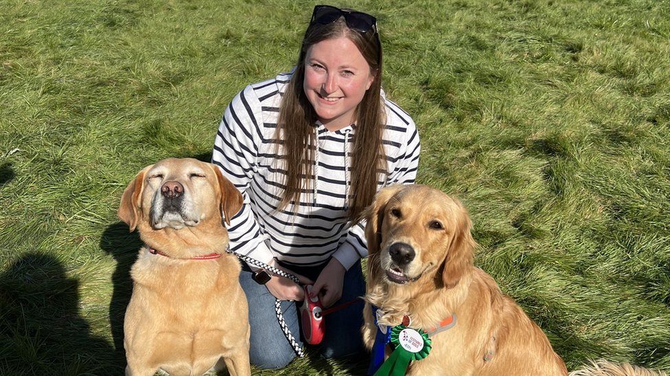 Olivia Riley with two dogs crouching down on grass.