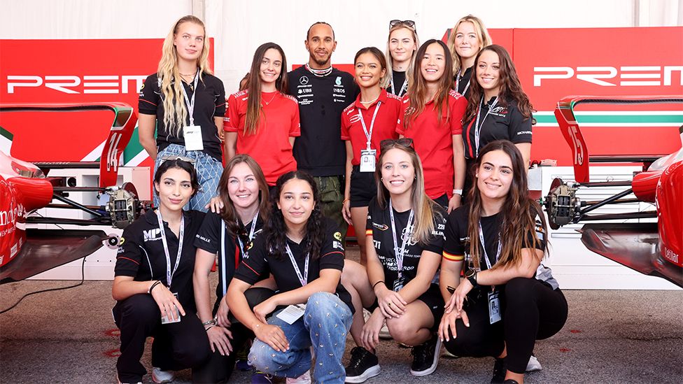 Female F1 Academy drivers with Lewis Hamilton who is standing in the middle, wearing a black top with branding. The others are wearing black and red tops with five of them kneeling in front as the others stand. The background has the rear wing of racing cars on each side, with red adversiting boards behind them.