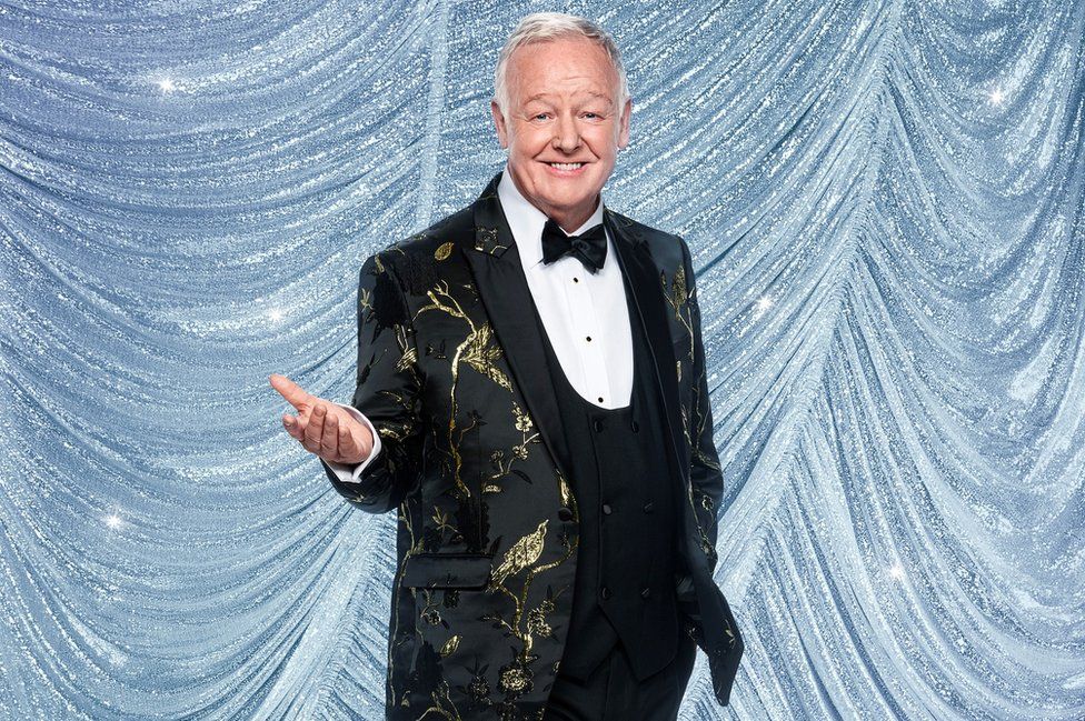 Les Dennis with his palm face up wearing a black and gold jacket