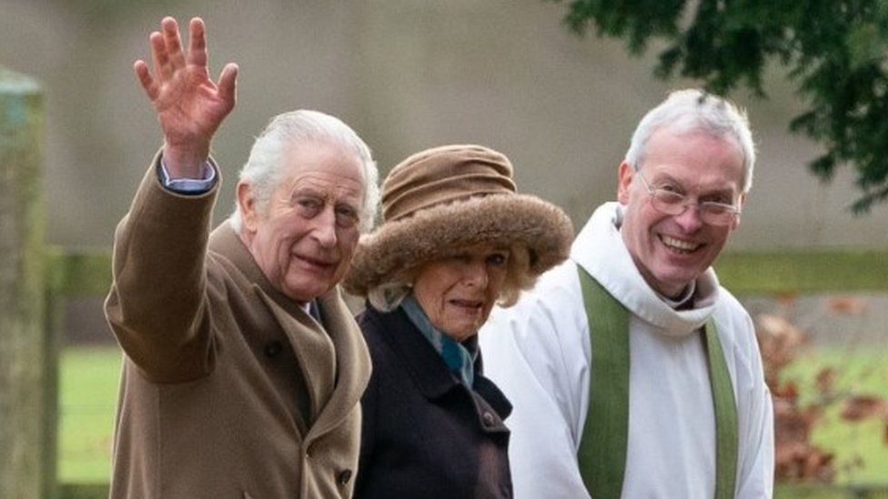 King Charles III waves to the cameras as he walks beside Queen Camilla