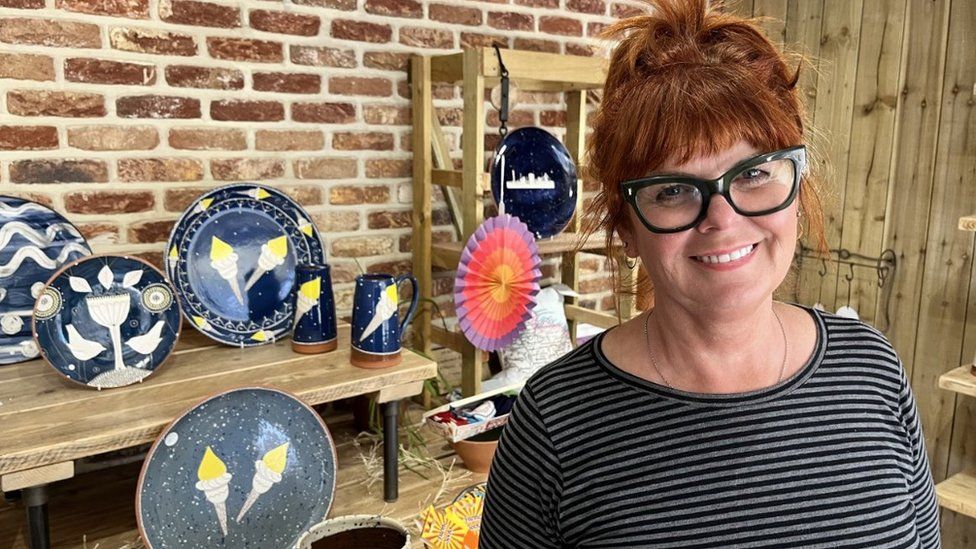 A woman in glasses smiles, standing in front of plates and pottery
