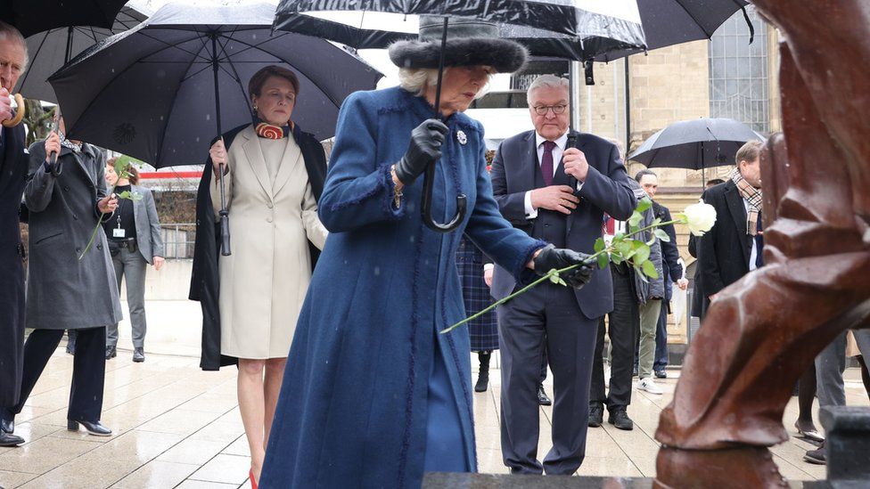 The Queen Consort lays a single white rose in a hat, blue jacket and dress and brooch. She is covered by an umbrella