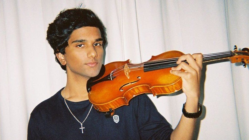 Joel Sunny holds his violin and looks into the camera. He stands in front of a white curtain and wears a black long sleeve sweater and a cross chain around his neck