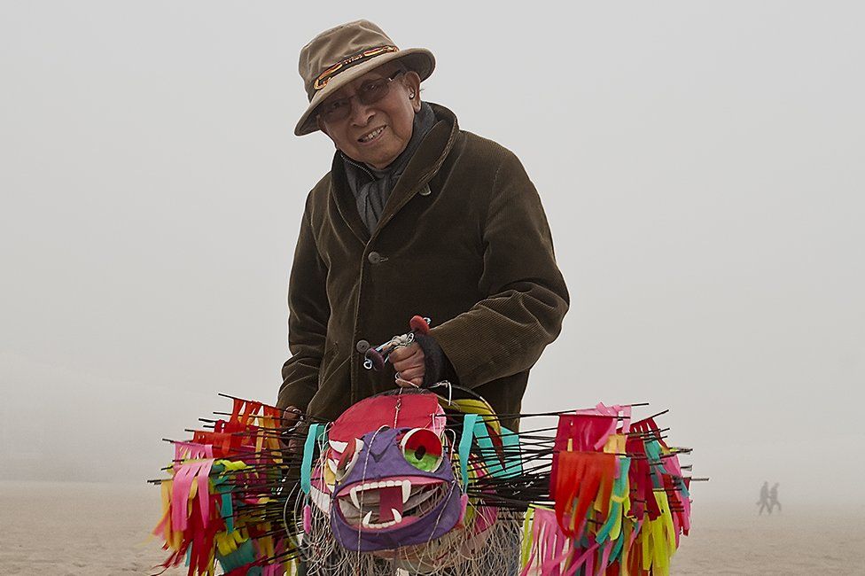 A photograph shows Tyrus Wong holding one of his intricately made bamboo kites on the beach