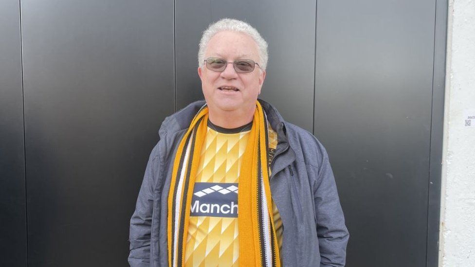 An older white man with white hair and tinted glasses smiles at the camera in a bright yellow Maidstone shirt, scarf and grey coat