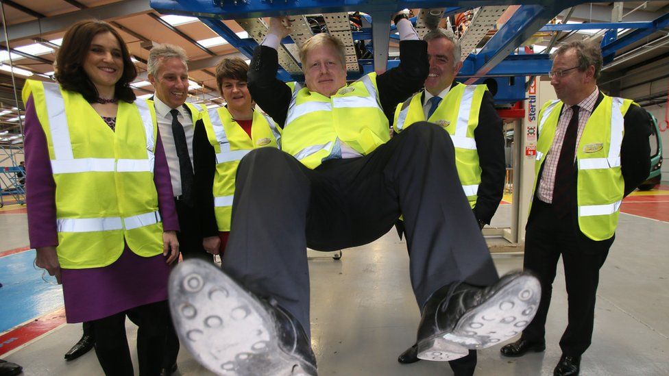 Boris Johnson swinging from a bus chassis in the Wrightbus factory while surrounded by other politicians