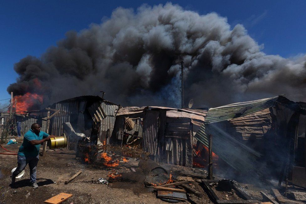 A resident throws water from a bucket as a fire rages through shacks in Masiphumelele, Cape Town, South Africa, 21 November 2022