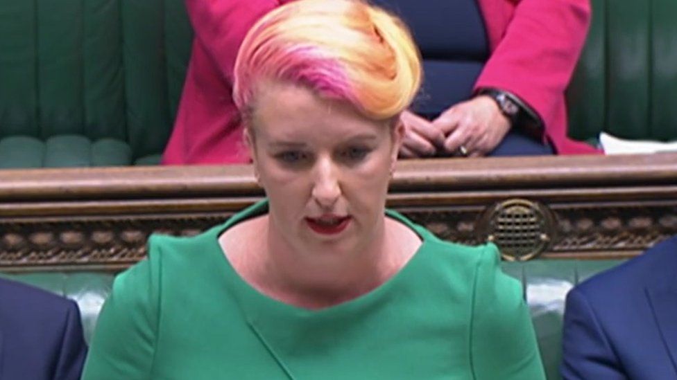 Woman with orange and pink-tinted hair wearing green top stands at opposition dispatch box