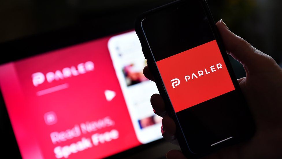 A photo illustration shows someone holding a phone with the Parler logo on it, while a laptop screen with its advertising is seen blurred in soft focus in the background
