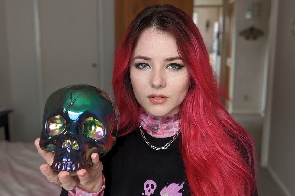 A woman with long, red/pink hair sits on a bed, holding an iridescent skull in her upturned left palm. She's giving a piercing stare to the camera, and wears a black top with pink-pattered high neck. She's got a chain-style silver necklace on too.