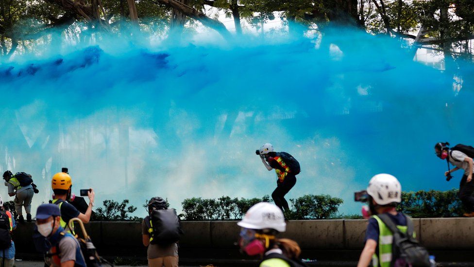 A wave of surreal blue water streaking through the air as journalists and protesters duck to avoid the dye in an open space