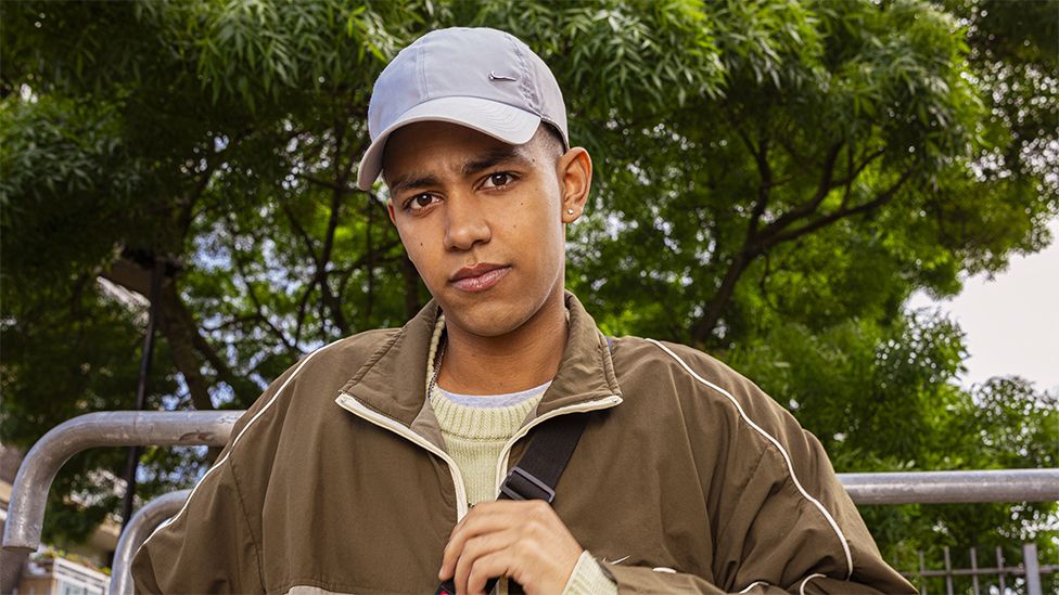 Character of Junior wearing a brown jacket and cream sweater and grey cap. He is looking at the camera while clucthing the top of a bag strap running across his chest. The background is of green trees.