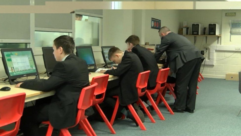 Four pupils sitting in front of a row desks with computer screens and a teacher helping one student at the end of the row