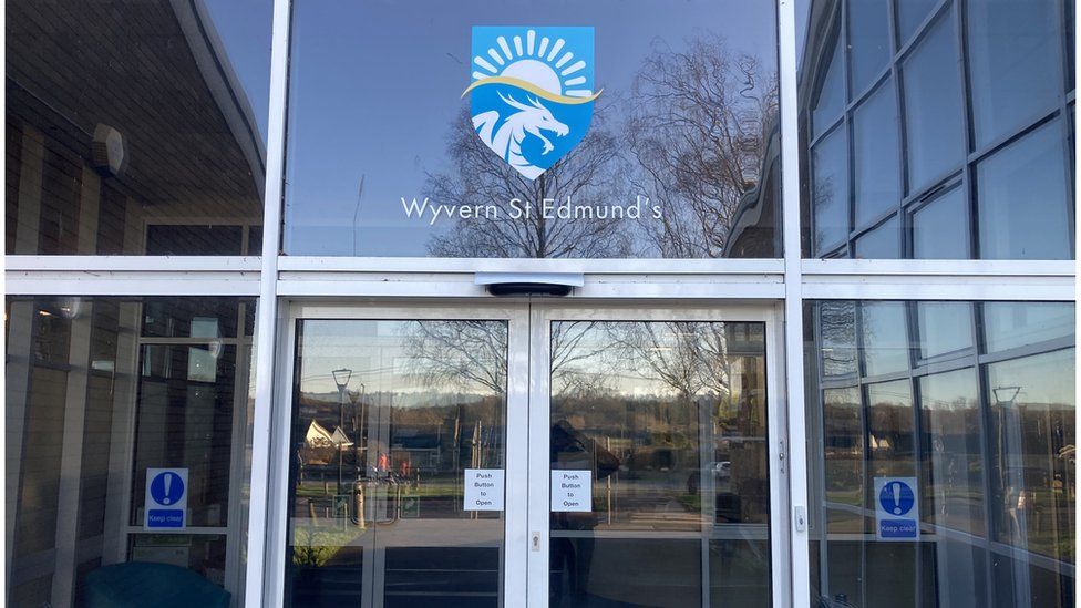 The front doors of the school - large and glass with the logo above, which includes the wyvern creature, which looks a big like a dragon