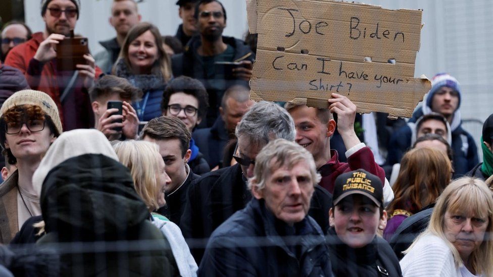 A man in a crowd holds a sign that reads: Joe Biden, can I have your shirt?