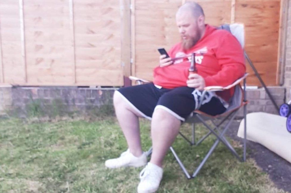 A large man in a red hoody and shorts sits in a garden chair