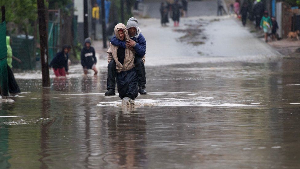 Мужчина несет пожилого мужчину через затопленную дорогу во время сильного наводнения в результате шторма в деревне сэра Лоури недалеко от Сомерсет-Уэста, 25 сентября 2023 года.