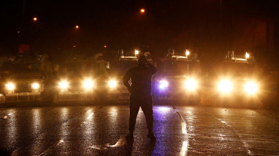 A man stands facing a line of police armoured Land Rovers