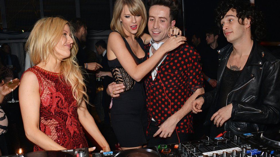 Ellie Goulding, Taylor Swift, Nick Grimshaw and Matty Healy stand around a set of DJ decks at an awards party in celebratory scenes