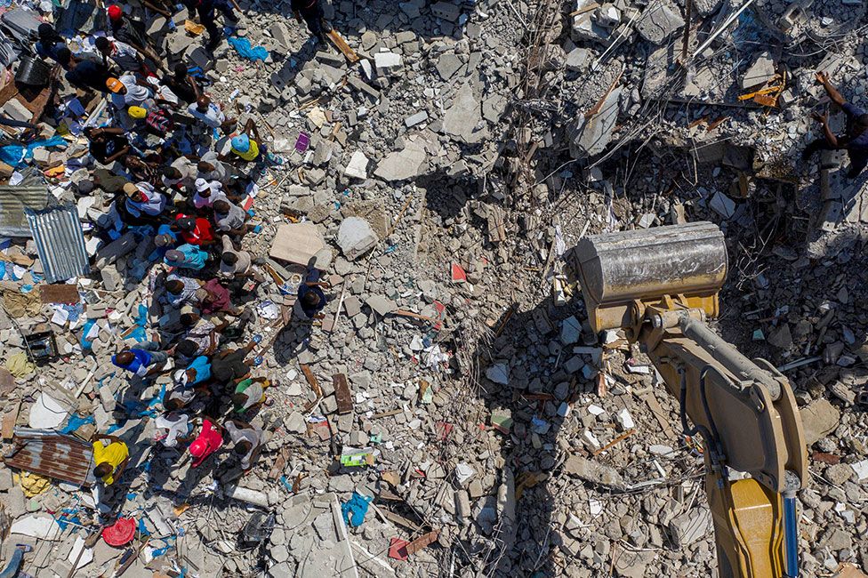 People watch an excavator removing rubble from the site of a collapsed hotel in Les Cayes, Haiti, on 16 August 2021