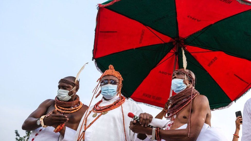 Oba of Benin being led into the palace with two men supporting him at his side. All three men are under an umbrella and they are wearing masks and traditional clothing with traditional beads. The Oba is wearing sunglasses.