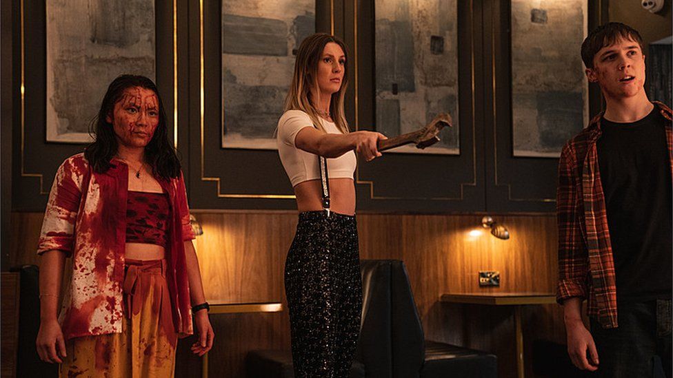 L-R Thaddea Graham as Vivian, Miya Ocego as Rosie and Oscar Kennedy as Jamie in Wreck series one. Vivian, who has dark shoulder length hair and wears an open white shirt over a crop top and yellow trousers is covered in blood. Rosie stands about a foot away from her, her long ombre hair worn loose over her outfit of a white crop top and sparkly black trousers held up with silver braces. In her right hand she holds a large spanner outstretched in front of her towards someone out of shot. Jamie, a young white man with short brown hair, stands on the left wearing a red lumber-jack style shirt over a black T-shirt. He has blood on his face and looks off camera with a repulsed expression. They are inside on the cruise ship in what appears to be a restaurant, with brown panelled walls with a gold inlaid trim and four matching blue and white artworks hanging above lit tables behind them.