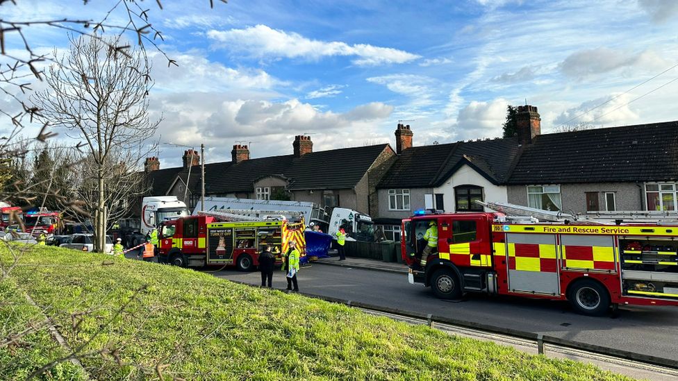 White lorry with smashed windscreen appears to be in contact with a row of houses after apparently leaving the road. Firefighters and police in hi-vis stand in the foreground on the road, with several fire engines visible