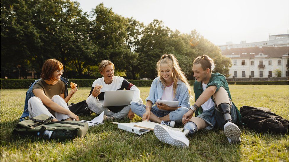 A group of university students sit on a field looking at books and laptops