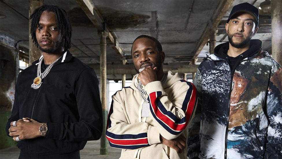 Krept and Konan and DJ Target, standing next to one another looking at the camera.