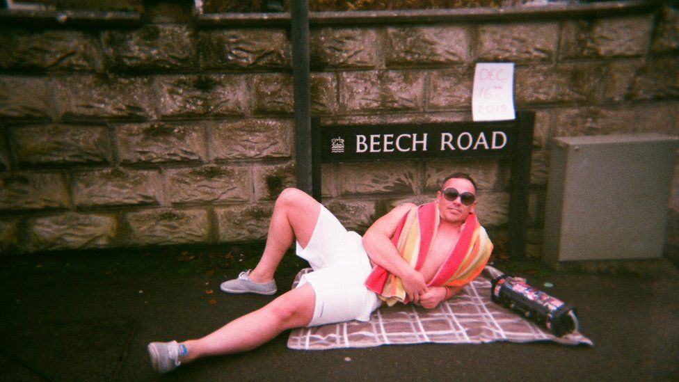 A man wearing shorts, sunglasses and a towel around his shoulders lies on a blanket on the floor in front of a street sign