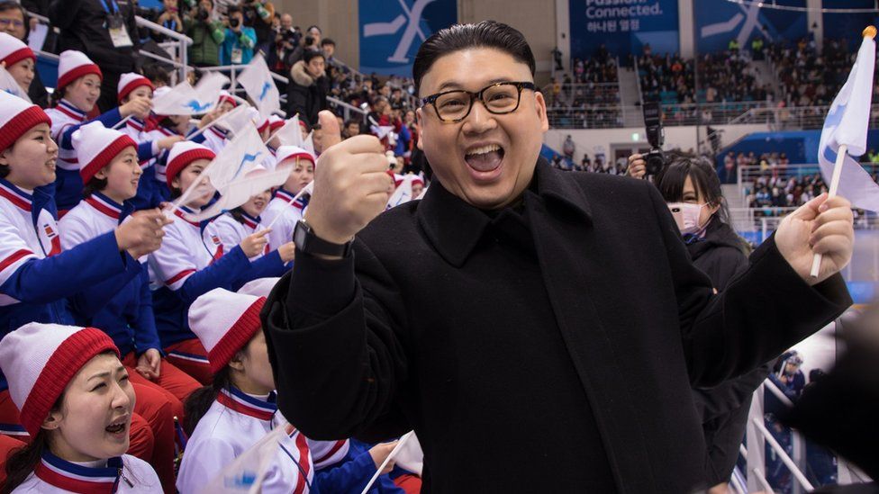 A Kim Jong-un impersonator gives a thumbs-up to the camera as Korea fans look on angrily