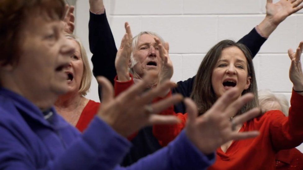 A photograph of former Welsh rugby player JPR Williams singing with choir The Forget-Me-Not-Chorus. People in the photo have their arms outstretched and mouths wide open
