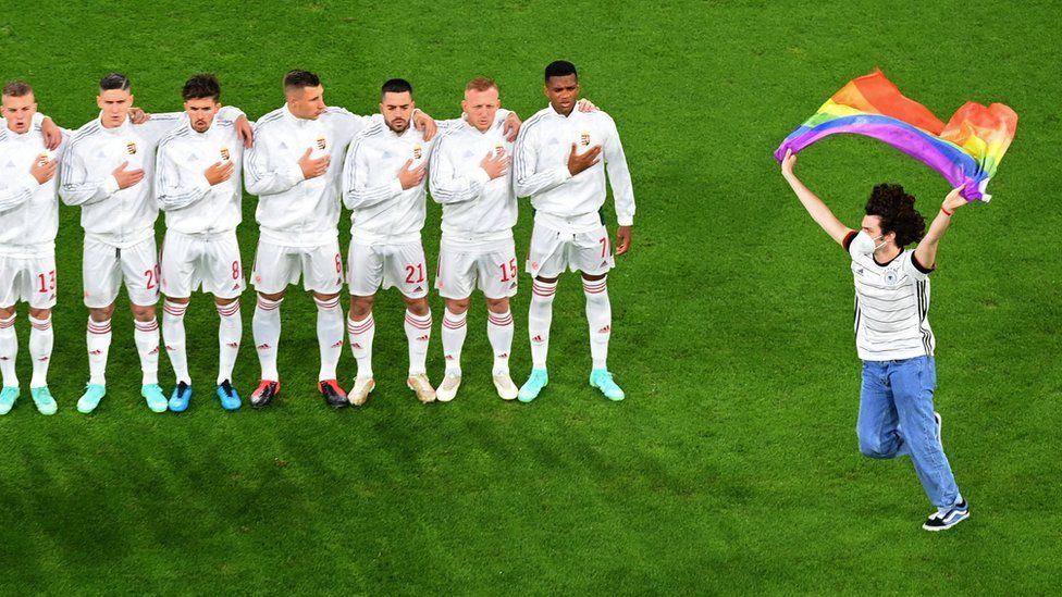 A person waving the rainbow flag protests next to Hungary team during national anthems at Germany v Hungary match in Munich, 23 Jun 21