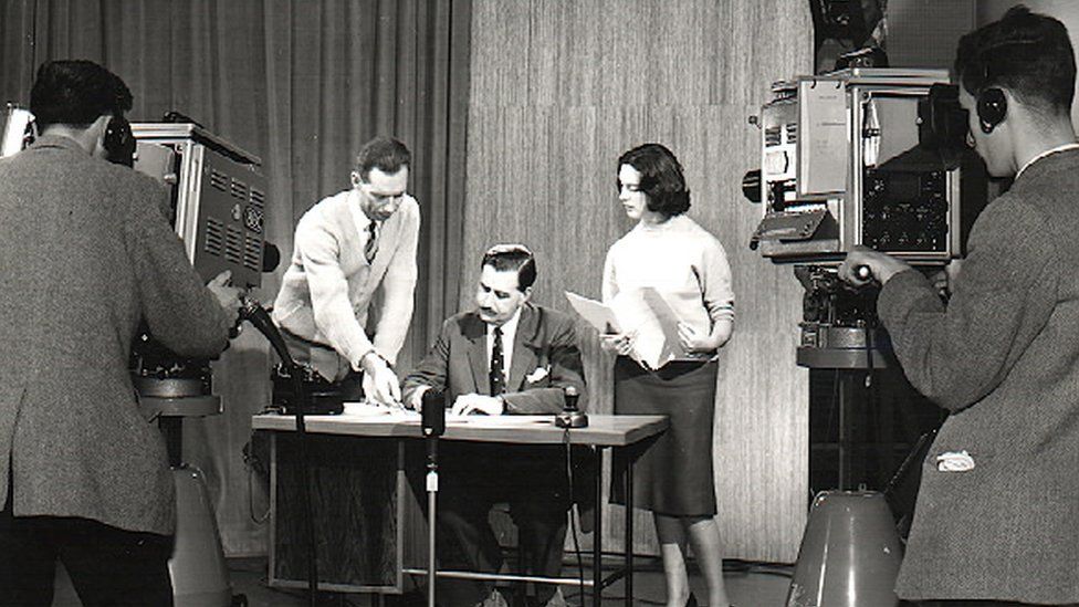 A black and white image in which Cecil Taylor leans across a desk to point out something to a newsreader