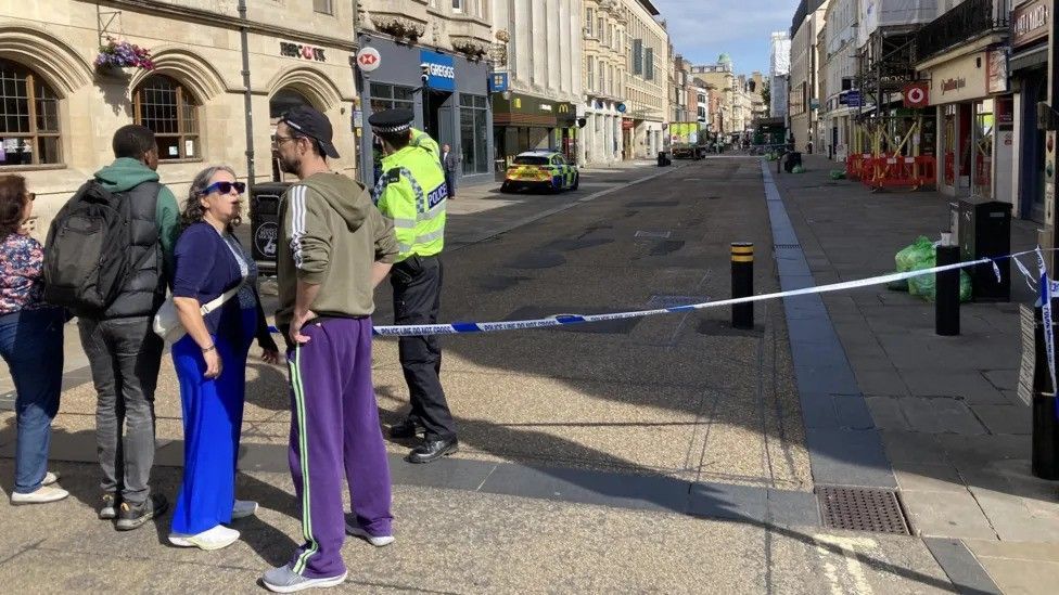 Bystanders and onlookers gather at a cordon on Cornmarket Street. A police car is in the distance outside Barclays bank.