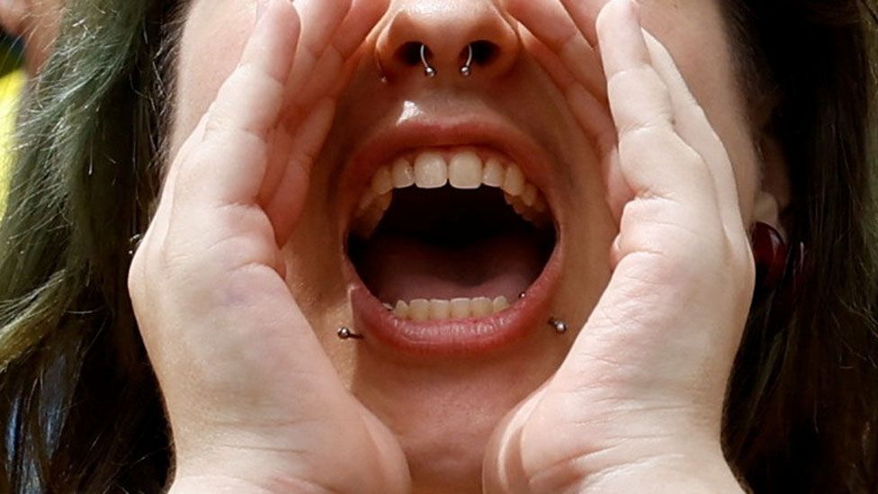 A woman with nose and lip piercings cupping her mouth with her hands while shouting