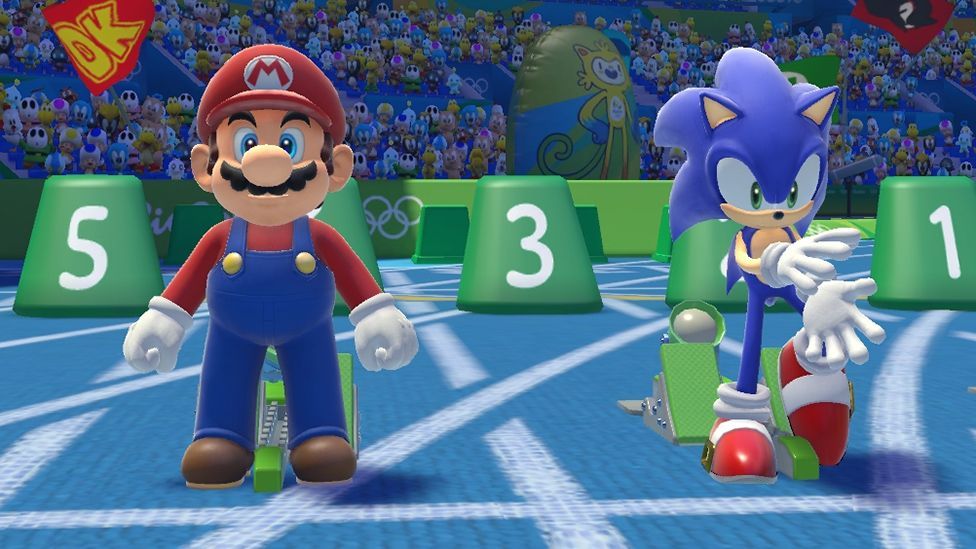 A screenshot showing Mario and Sonic at the starting blocks on a light blue racetrack. Mario wears his trademark dungarees and Sonic his famous red sneakers. The two are getting ready to race, with a crowd made up of Nintendo and Sega sidekick characters looking on behind them.