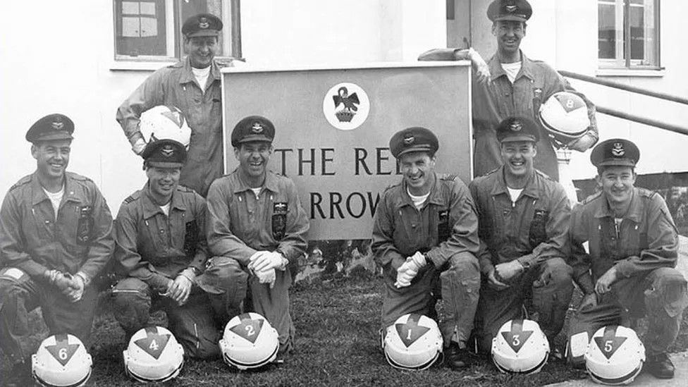 Black and white photograph showing members of the original team posing in front of a sign and building with their numbered helmets