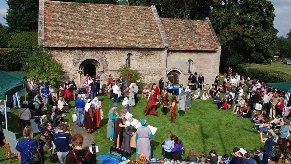 A group of medieval re-enactors, some dancing, others singing in front of an audience watching them, outside a medieval stone chapel, Cambridge 