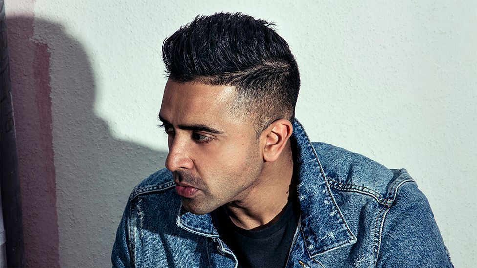 Jay Sean, a man looking to his right, wearing a blue denim top and black tshirt.