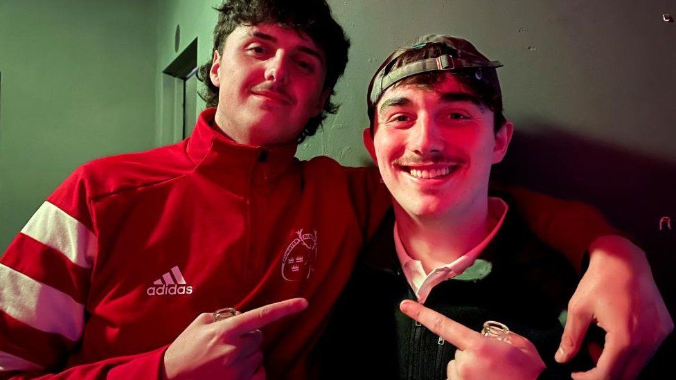 Two young men pointing at each other. One wears a baseball cap. The other is in a red Adidas fleece.