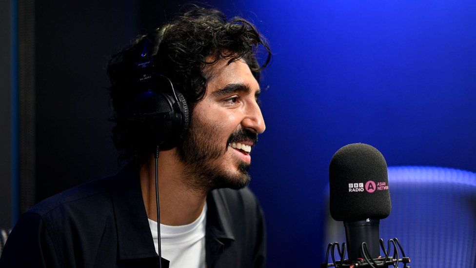 Dev Patel in the BBC Asian Network studio. Dev is a 33-year-old British Asian man with dark curly hair, a moustache and short beard and brown eyes. He's photographed from the side, smiling. He is pictured inside the studio, which has blue walls, and wears headphones as he sits in front of a microphone. He wears an unbuttoned black shirt over a white T-shirt