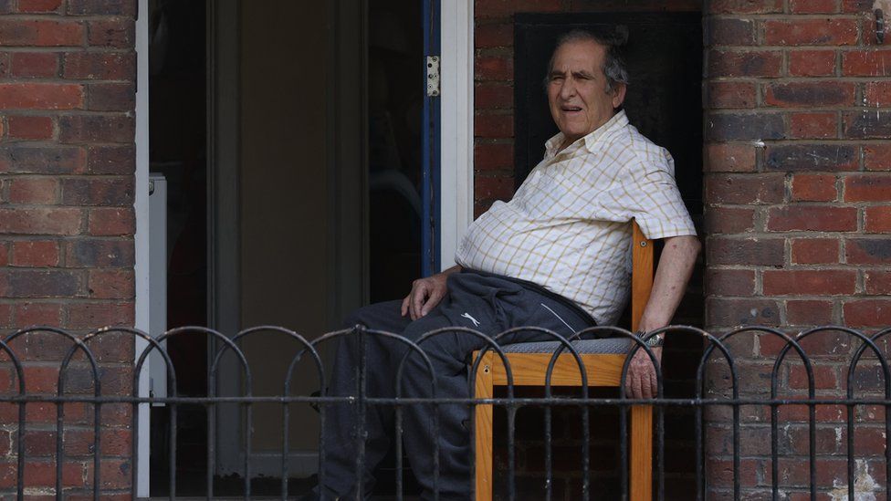 Пожилой мужчина сидит на деревянном стуле перед своим кирпичным зданием во время сильной жары в 2022 году