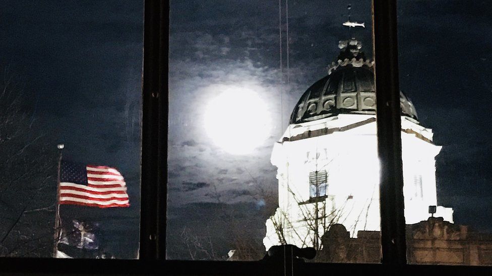 The moon appears bright white between a United States of America flag and the Indiana capitol building